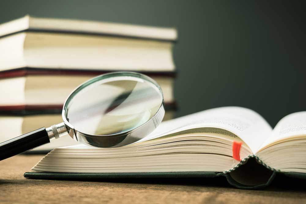 Magnifying glass on the opened book on the table, with pile of books on background.